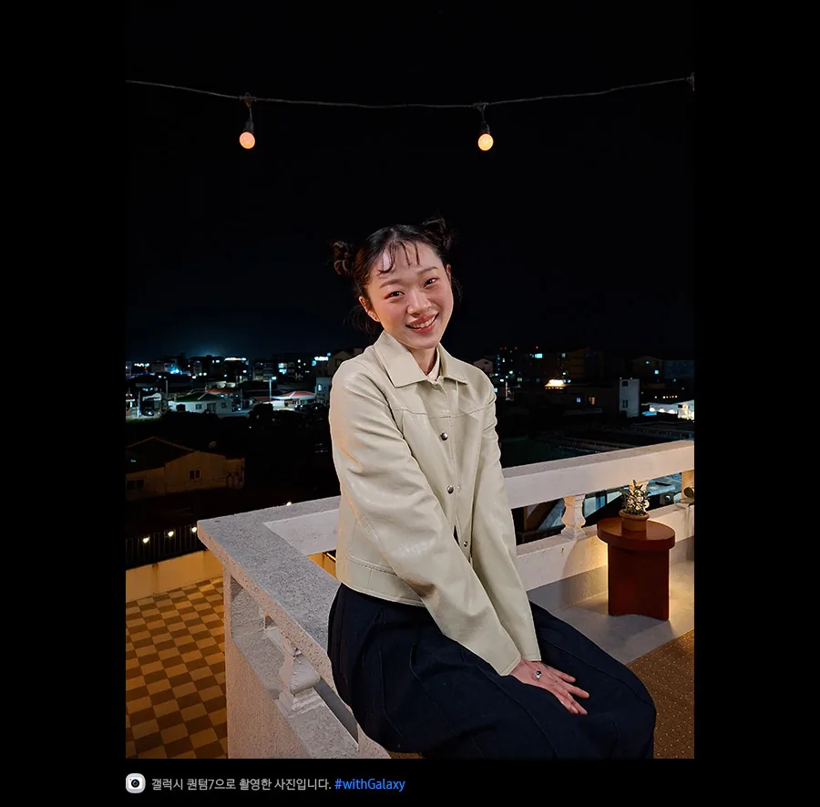 갤럭시 퀀텀7으로 촬영한 밤에 야외 테라스에 기대어 있는 젊은 여성의 사진입니다. 최적화된 나이토그래피 덕분에 사진의 디테일이 밝고 선명합니다. 하단에는 카메라 아이콘과 함께 '갤럭시 퀀텀7으로 촬영한 영상입니다' 문구와 #with_galaxy 해시태그가 표시되어 있습니다.